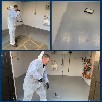 A person in protective coveralls painting a grey workshop floor with a roller.
